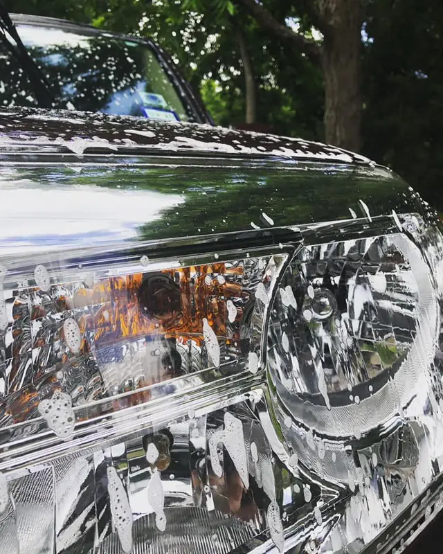 Soap suds covering the hood and headlight of a dark green SUV being washed outdoors under trees